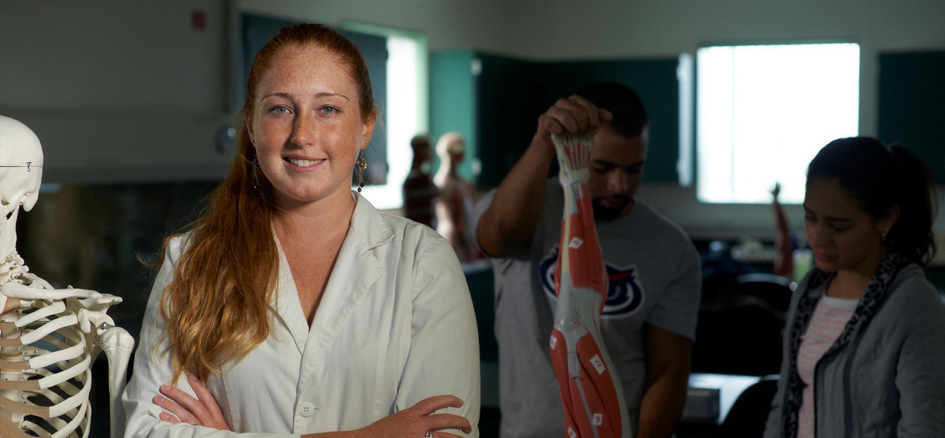 Nursing student in lab coat standing in classroom with students in background examining anatomical figure